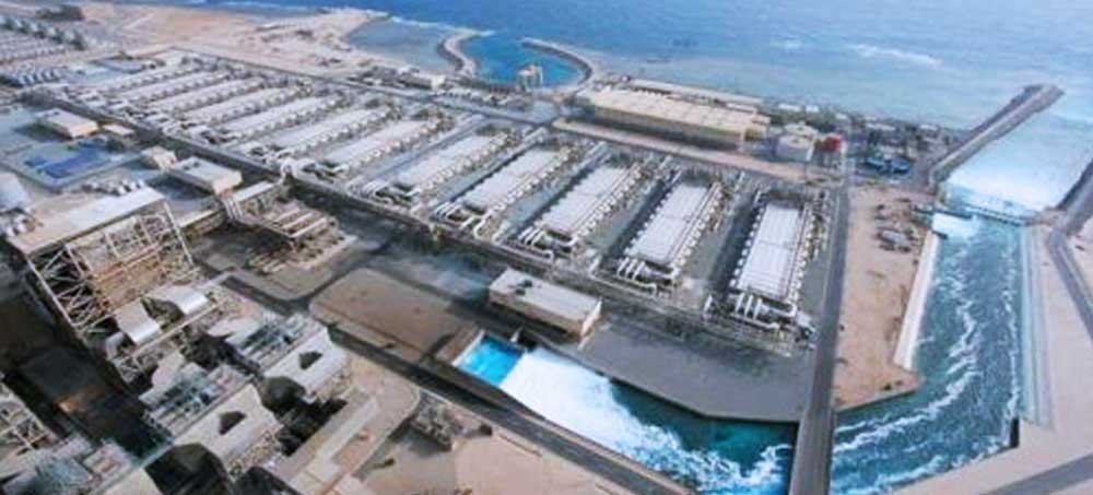 Aerial view of a large seawater desalination plant located on a coastal area, showing multiple industrial structures, pipelines, and water channels, with the ocean in the background.